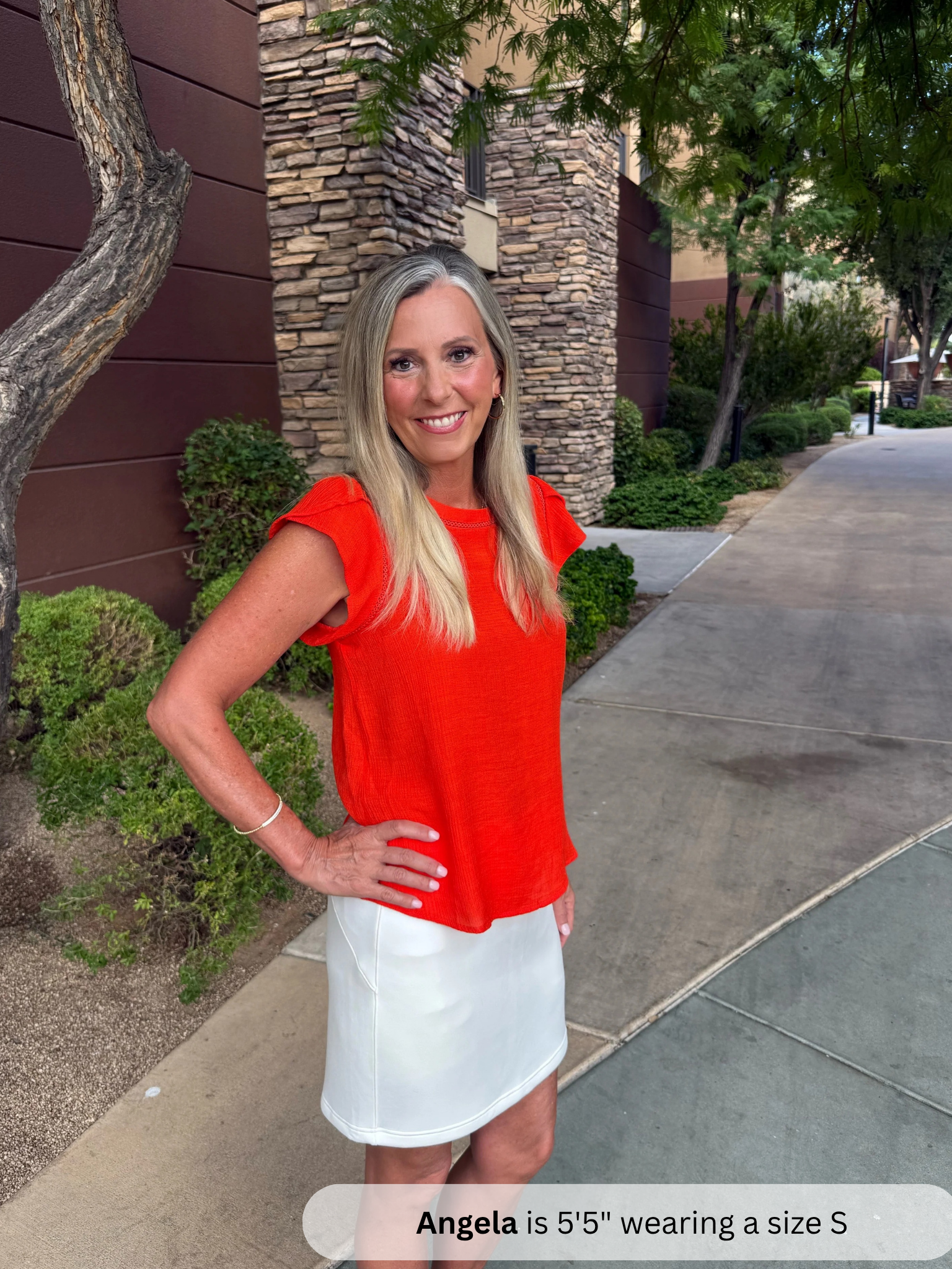 /Tomato Red/ in a red top and white skirt standing on a sidewalk with a stone building and trees in the background.
