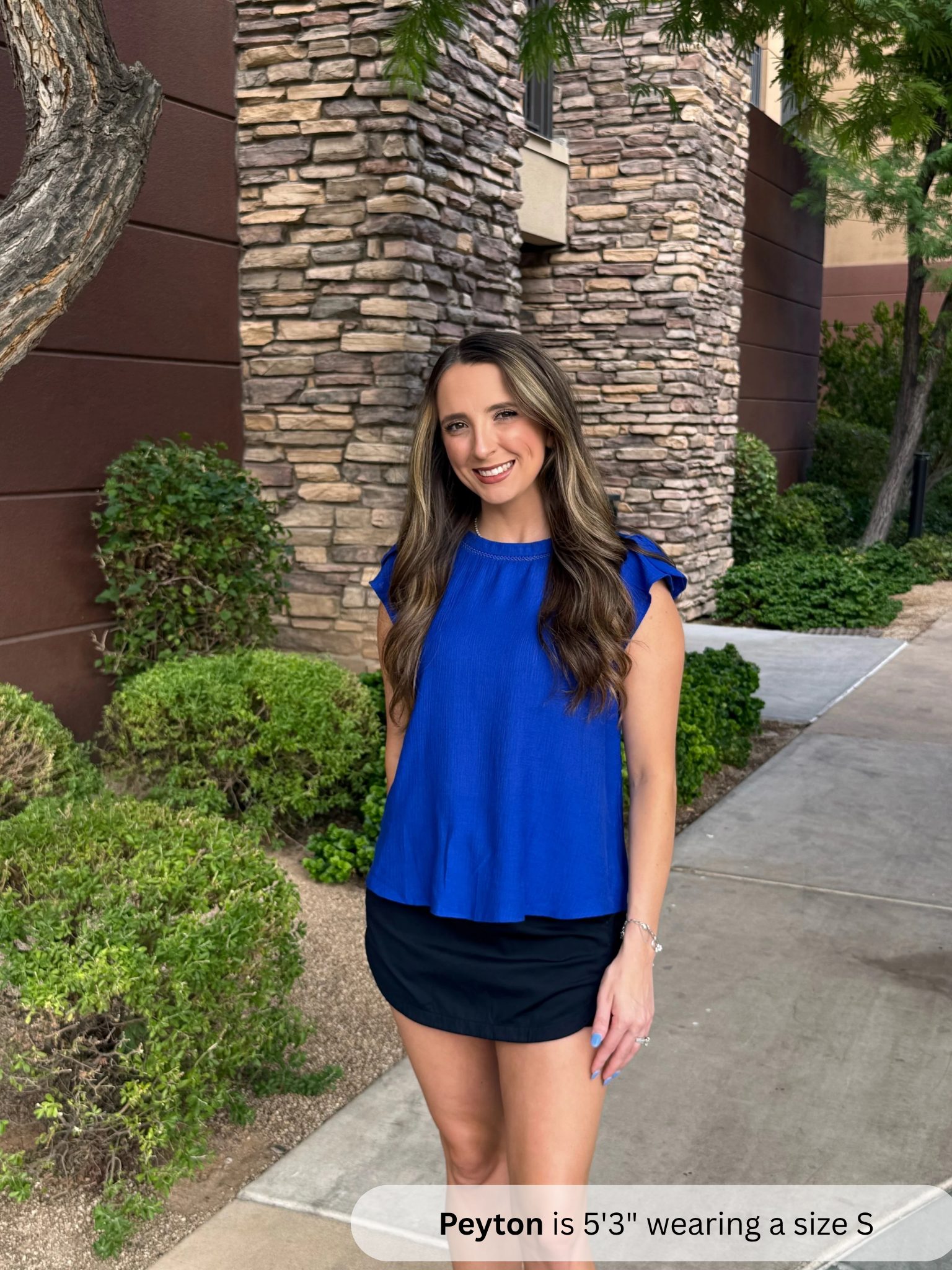 /Royal Blue/ Woman in a blue top and black skirt standing outdoors next to a stone wall.