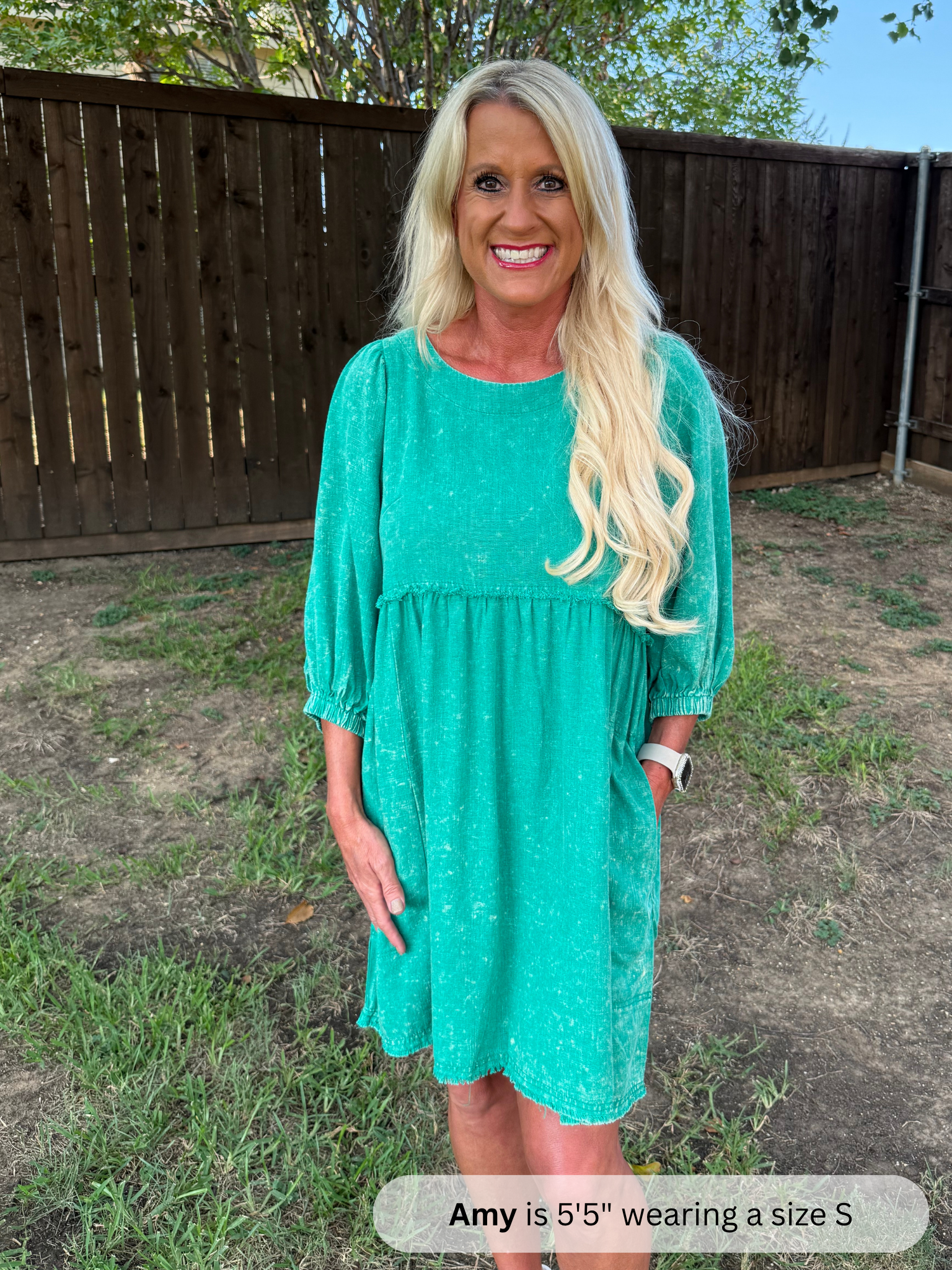 Woman wearing a teal dress standing outdoors with a wooden fence and grass in the background.