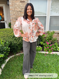 Woman standing outdoors in a garden wearing a floral blouse and gray pants.