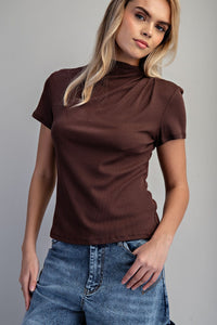 A woman wearing a short-sleeved brown ribbed turtleneck paired with classic blue denim jeans, photographed against a neutral gray background.