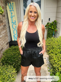 Woman wearing a black tank top and leather shorts standing in front of a house with a 'Welcome' sign.