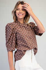 Woman wearing a brown polka dot blouse with puffed three-quarter sleeves and a tie front detail, paired with white pants, against a neutral background.