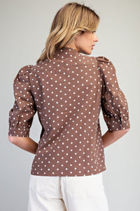A woman wearing a brown polka dot blouse with white dots and three-quarter length puffed sleeves, paired with white pants, shown from the back against a neutral gray background.