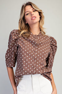 Woman wearing a brown polka dot blouse with puffed three-quarter sleeves and button-front closure, paired with white pants against a neutral background.