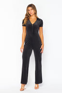 A woman wearing a black ribbed short-sleeve jumpsuit with a V-neckline and full-length straight legs, styled with metallic sandals against a white background.