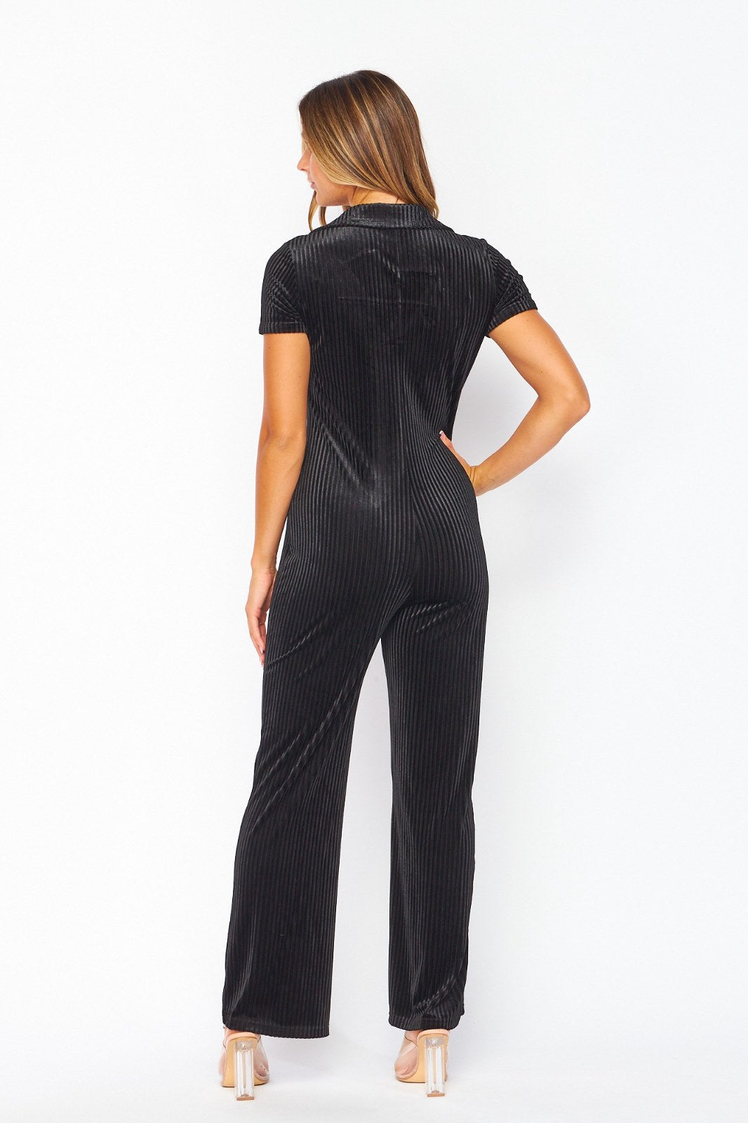 Woman wearing a black ribbed short-sleeve jumpsuit with a wide-leg pant silhouette, photographed from behind against a white background. The outfit features a fitted bodice and is styled with nude platform heels.