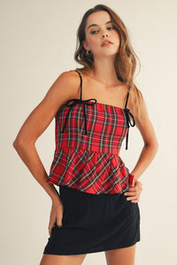 A woman wearing a red tartan plaid camisole top with thin black straps and a drawstring detail, paired with a black mini skirt. The top features a fitted bodice with a peplum ruffle at the waist.