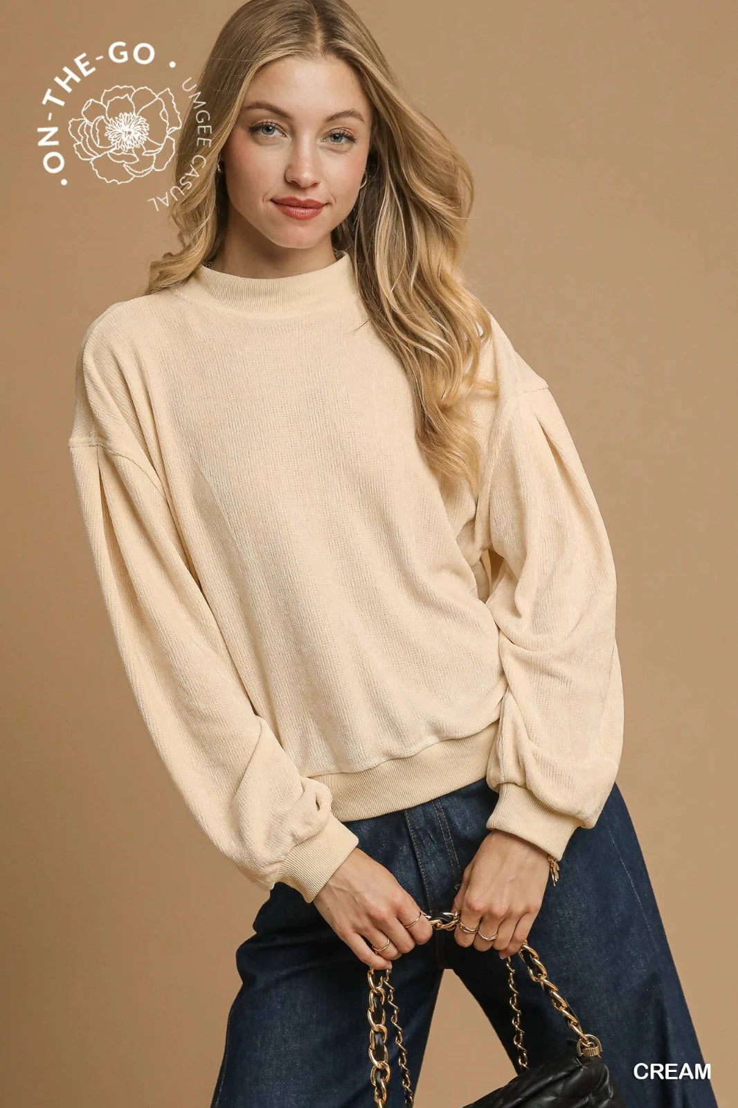 A woman wearing a cream-colored ribbed knit sweater with voluminous puffed sleeves and a cropped fit, paired with dark denim jeans and a chain-link shoulder bag against a tan background.