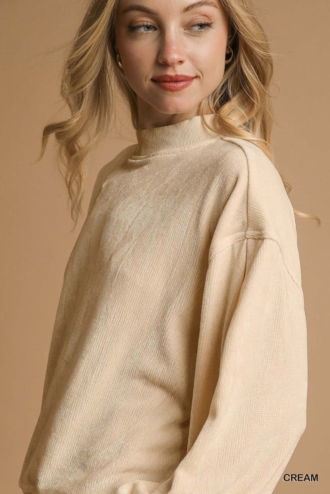 A woman wearing a cream-colored ribbed knit sweater with a mock turtleneck and long sleeves, posed against a neutral tan background.