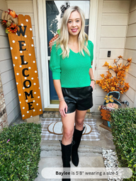 Woman standing in front of a house with Halloween decorations, wearing a green top and black leather shorts.