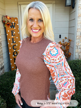 Woman wearing a brown sweater with colorful sleeves in front of a house with a 'Welcome' sign.