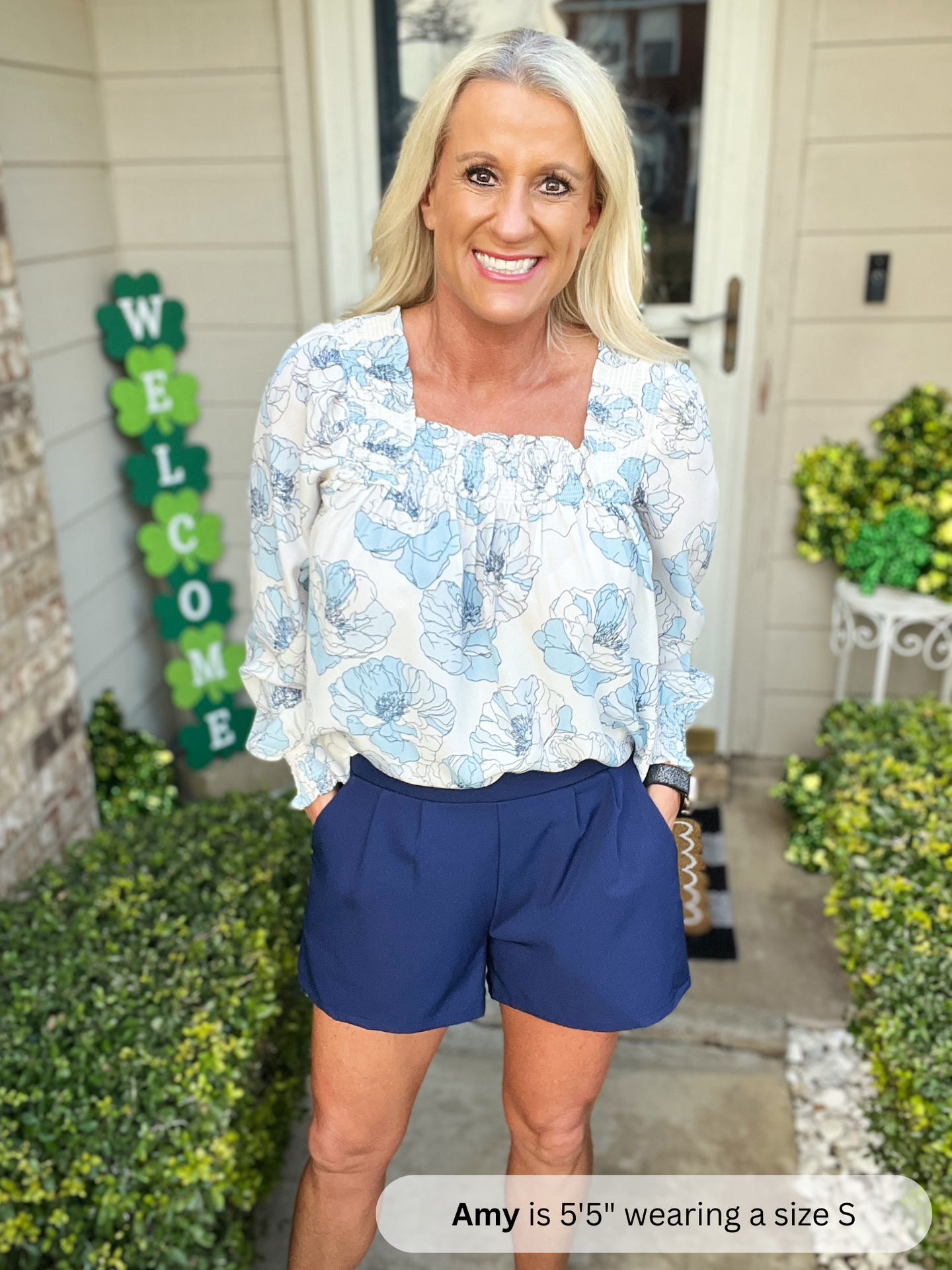 Woman standing on a porch wearing a floral blouse and blue shorts, with a 'Welcome' sign in the background.