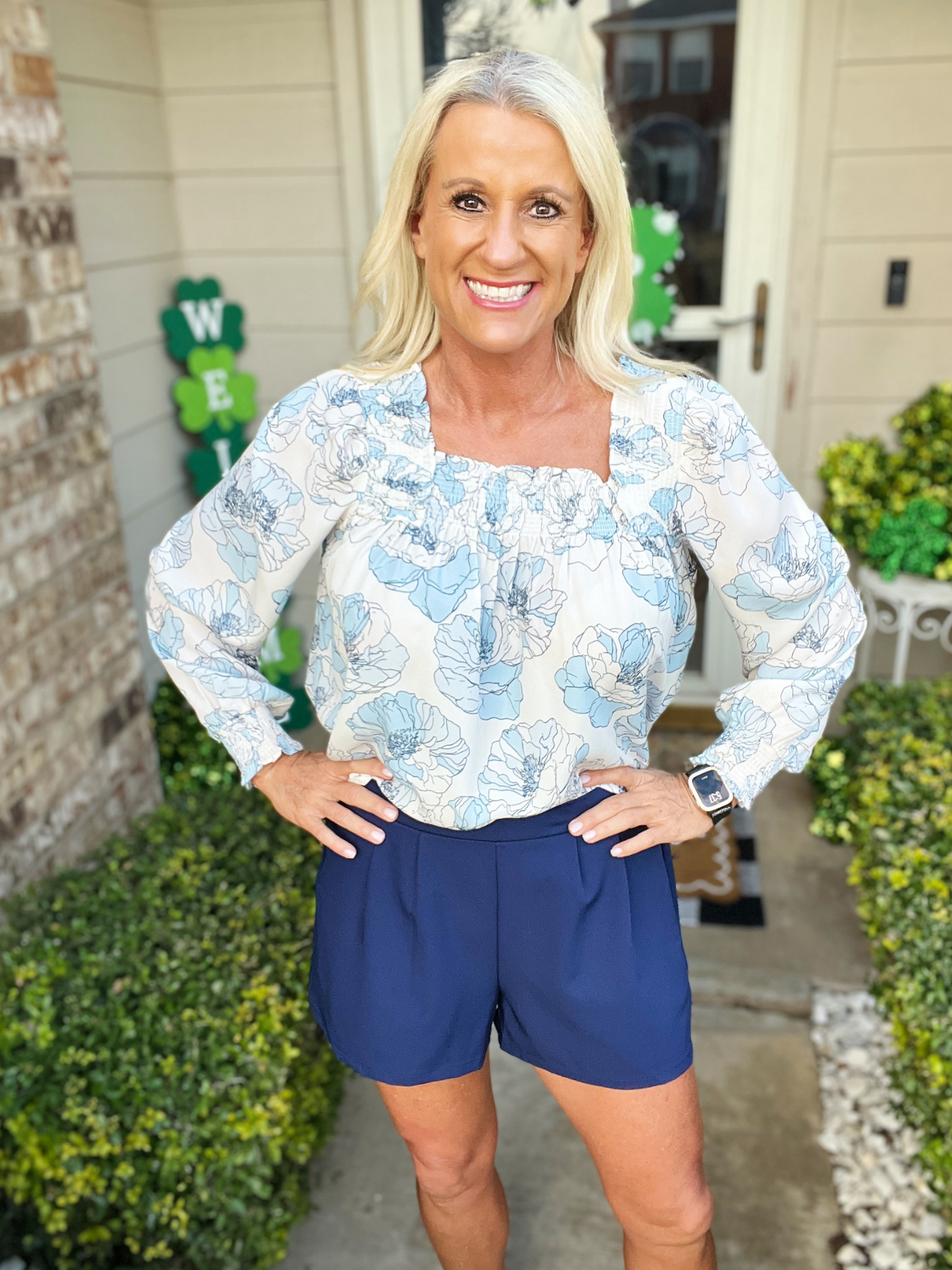 Woman wearing a floral blouse and blue shorts standing outdoors.