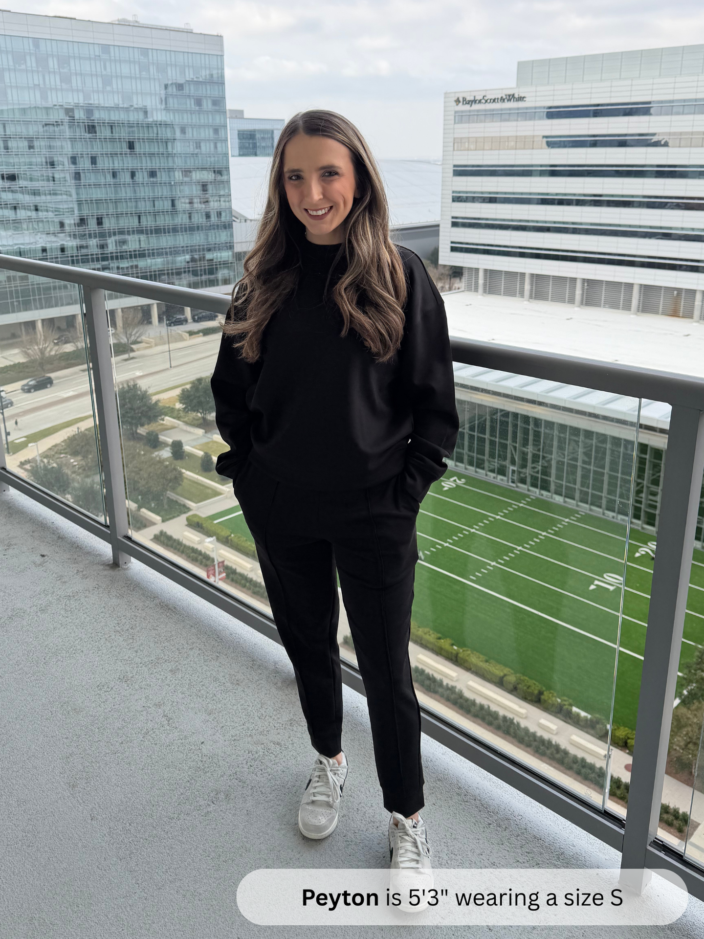 Person wearing a black outfit standing on a balcony with a view of a sports field and buildings.