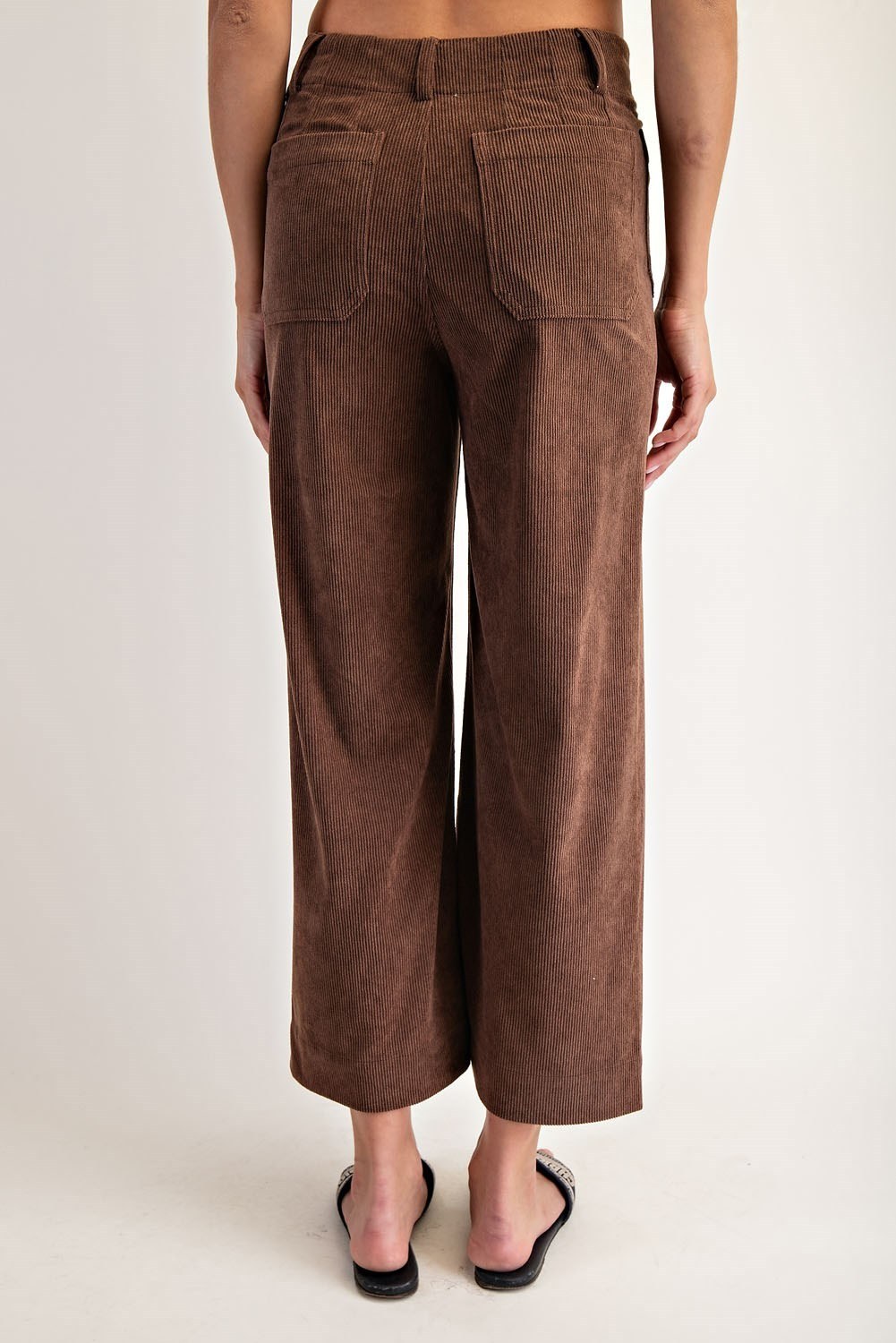 Back view of a woman wearing brown corduroy cropped pants with a high waist and straight leg silhouette, featuring visible seaming and functional pockets, paired with black platform heels.