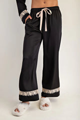 Black wide-leg lounge pants with an elastic waistband and cream drawstring, featuring decorative lace trim at the cuffs and waist. Modeled with matching black and cream fuzzy slippers.