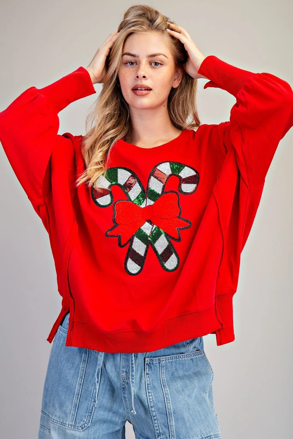 A woman wearing a bright red crew neck sweatshirt featuring a festive candy cane design with a red bow, paired with light blue jeans.