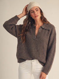 A woman wearing a charcoal gray ribbed knit sweater with studded collar detail and long sleeves, paired with white pants and a cream-colored headband.