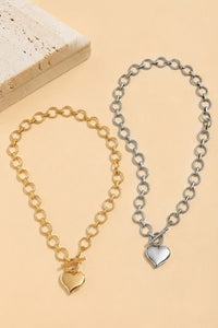 Two chain link bracelets displayed on a beige background: one in gold tone with a gold heart charm, and one in silver tone with a silver heart charm, both featuring rolo-style chain links.