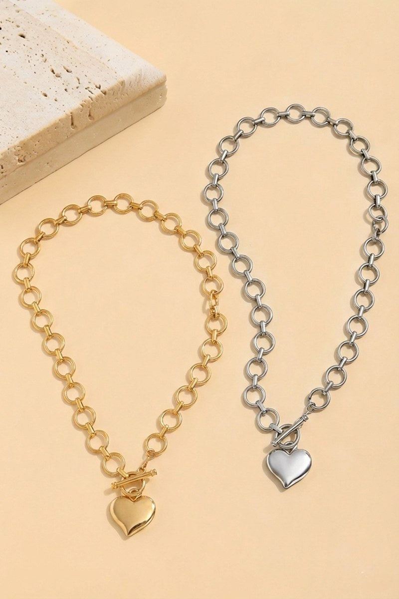 Two chain link bracelets displayed on a beige background: one in gold tone with a gold heart charm, and one in silver tone with a silver heart charm, both featuring rolo-style chain links.