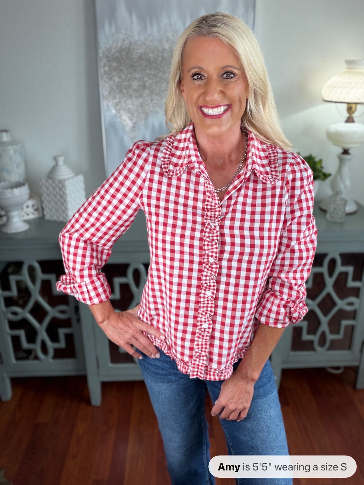 A woman wearing a red and white gingham check button-up shirt with ruffled placket and three-quarter sleeves, paired with blue jeans, standing in an indoor home setting.
