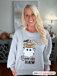 A woman wearing a light gray crewneck sweatshirt featuring a playful Halloween design with a ghost wearing a gold cowboy hat, black bats, a rope lasso, pumpkins, and the text "Boo HAW" in black embroidery.