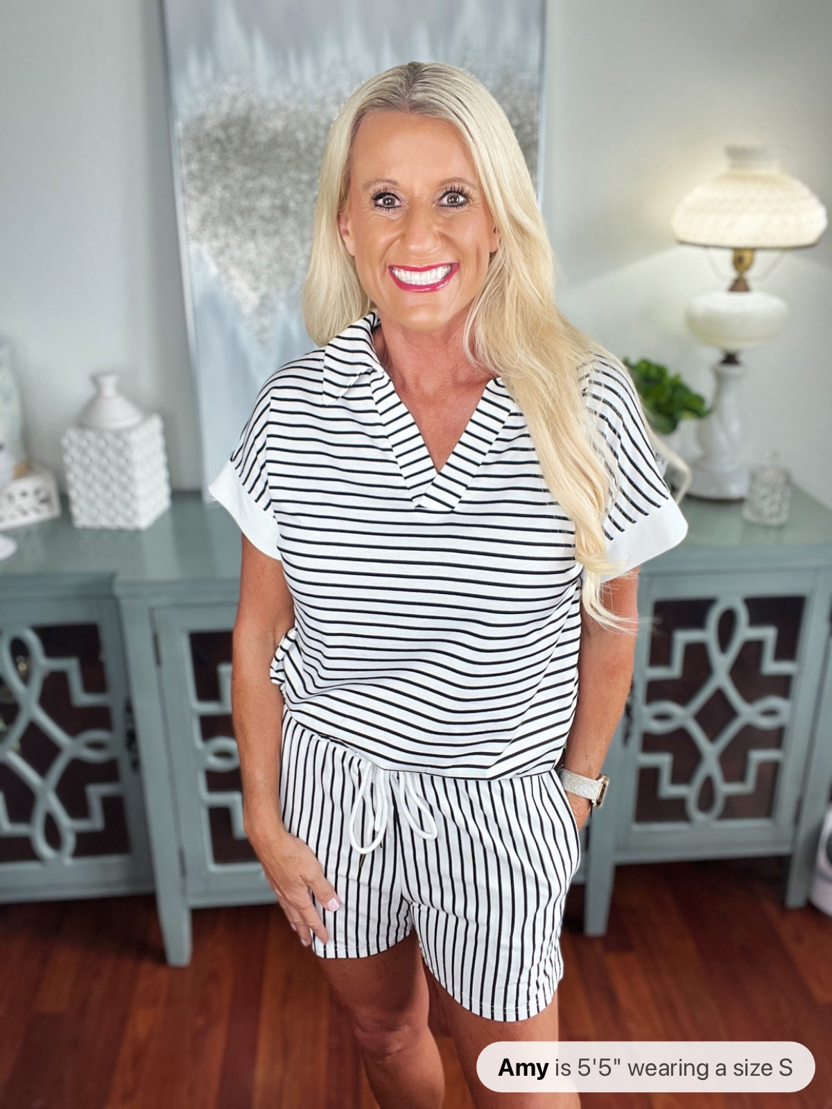 Woman wearing a white and navy striped short-sleeve shirt and matching shorts set, styled casually in a home interior setting with light blue cabinetry in the background.