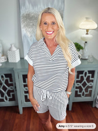 Woman wearing a white and navy striped short-sleeve shirt and matching shorts set, styled casually in a home interior setting with light blue cabinetry in the background.