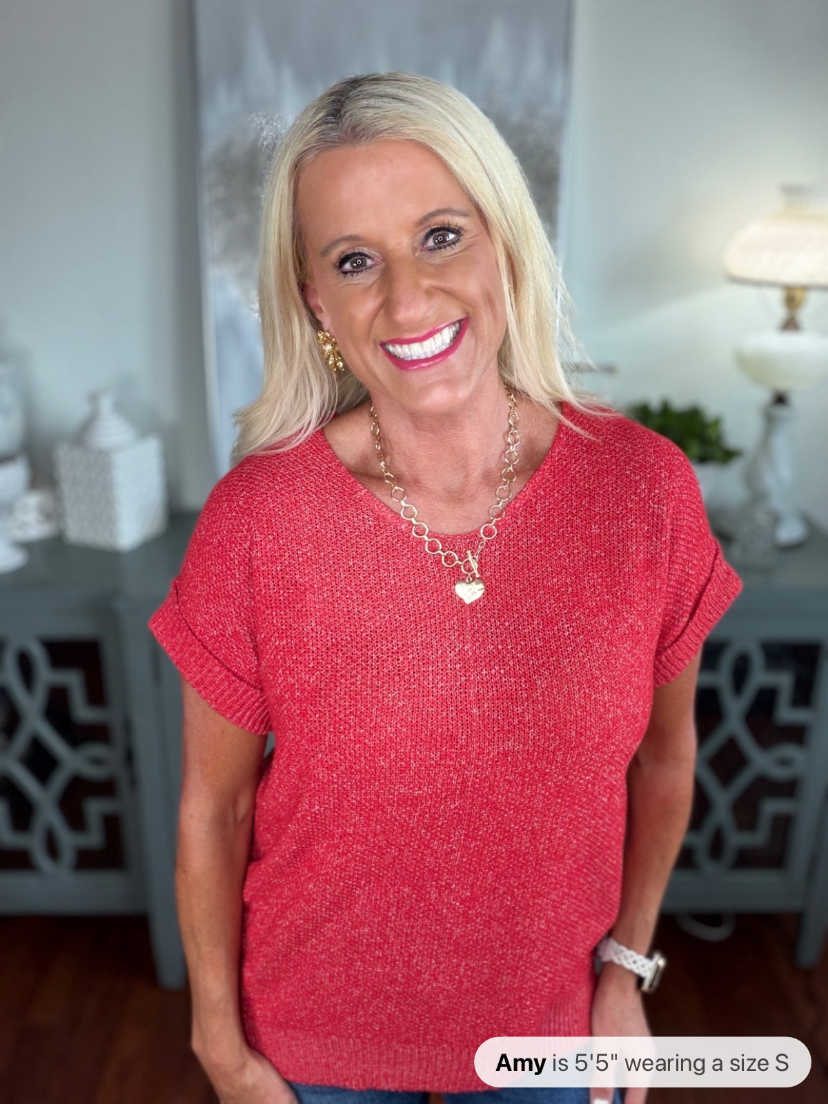 Woman wearing a short-sleeved red knit sweater with a crew neckline and rolled cuffs, styled with a silver pendant necklace and jeans.