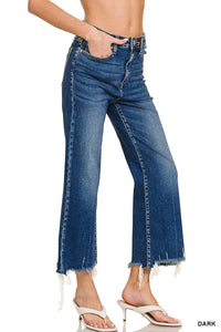 A woman wearing dark blue cropped jeans with a high waist, straight leg, and distressed frayed hem, paired with white slide sandals. The jeans feature classic five-pocket styling and visible stitching detail.
