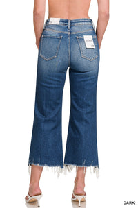 Rear view of a woman wearing dark blue cropped flare jeans with a high waist, featuring distressed frayed hems at the ankles and a white brand label on the back pocket.