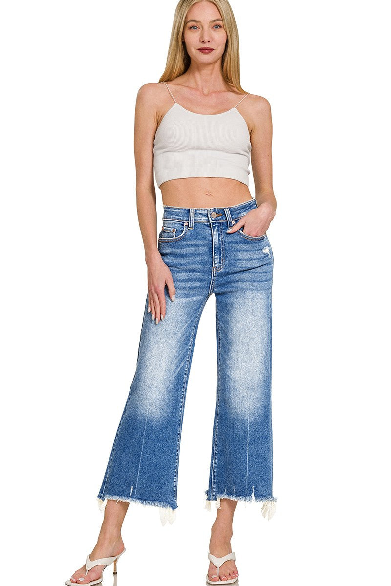 A woman wearing a white ribbed crop tank top paired with medium-wash blue jeans featuring a high waist, distressed detailing, and frayed cropped hem, styled with white strappy heels.
