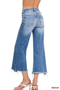 A woman wears medium-wash cropped wide-leg jeans with a high waist and frayed raw hem edges at the ankles. The jeans feature classic five-pocket styling and are paired with nude slide sandals.