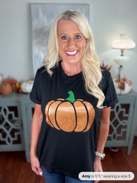 A woman wearing a black short-sleeve t-shirt featuring a large glittery orange pumpkin with a green stem on the front, styled with blue jeans in a bright, autumn-themed interior setting.