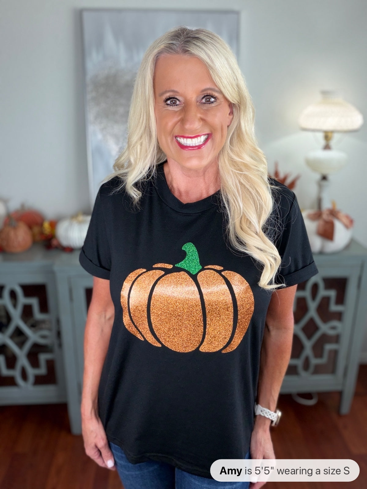 A woman wearing a black short-sleeve t-shirt featuring a large glittery orange pumpkin with a green stem on the front, styled with blue jeans in a bright, autumn-themed interior setting.