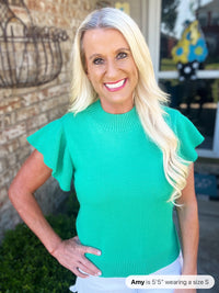 A woman wearing a turquoise ribbed knit sweater with short ruffled flutter sleeves and a crew neckline, paired with white pants.