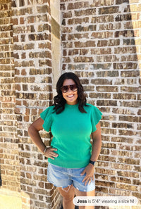 A woman wearing a bright turquoise short-sleeve knit sweater with a textured ribbed pattern, paired with distressed light blue denim shorts, poses against a brick wall.