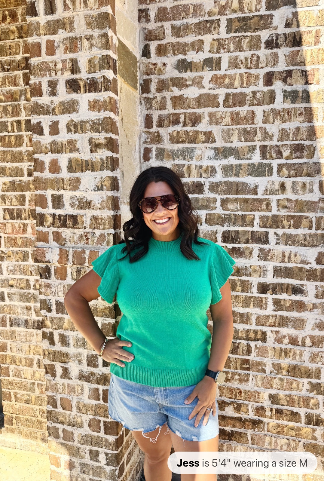 A woman wearing a bright turquoise short-sleeve knit sweater with a textured ribbed pattern, paired with distressed light blue denim shorts, poses against a brick wall.