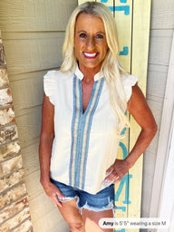 A woman wearing a white sleeveless blouse with blue geometric trim down the front and ruffled cap sleeves, paired with distressed denim shorts.