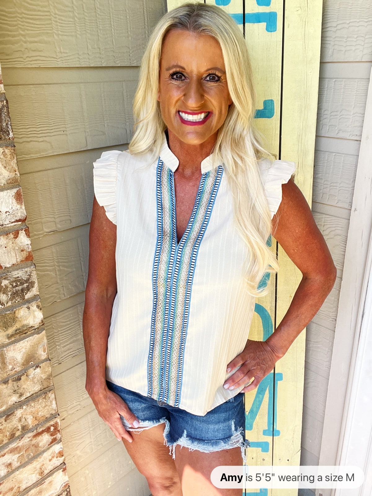 A woman wearing a white sleeveless blouse with blue geometric trim down the front and ruffled cap sleeves, paired with distressed denim shorts.