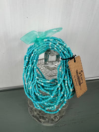 Turquoise beaded necklace with multiple strands of small seed beads arranged in a draped style, topped with a matching turquoise ribbon bow, displayed on a clear acrylic stand with a kraft paper price tag.