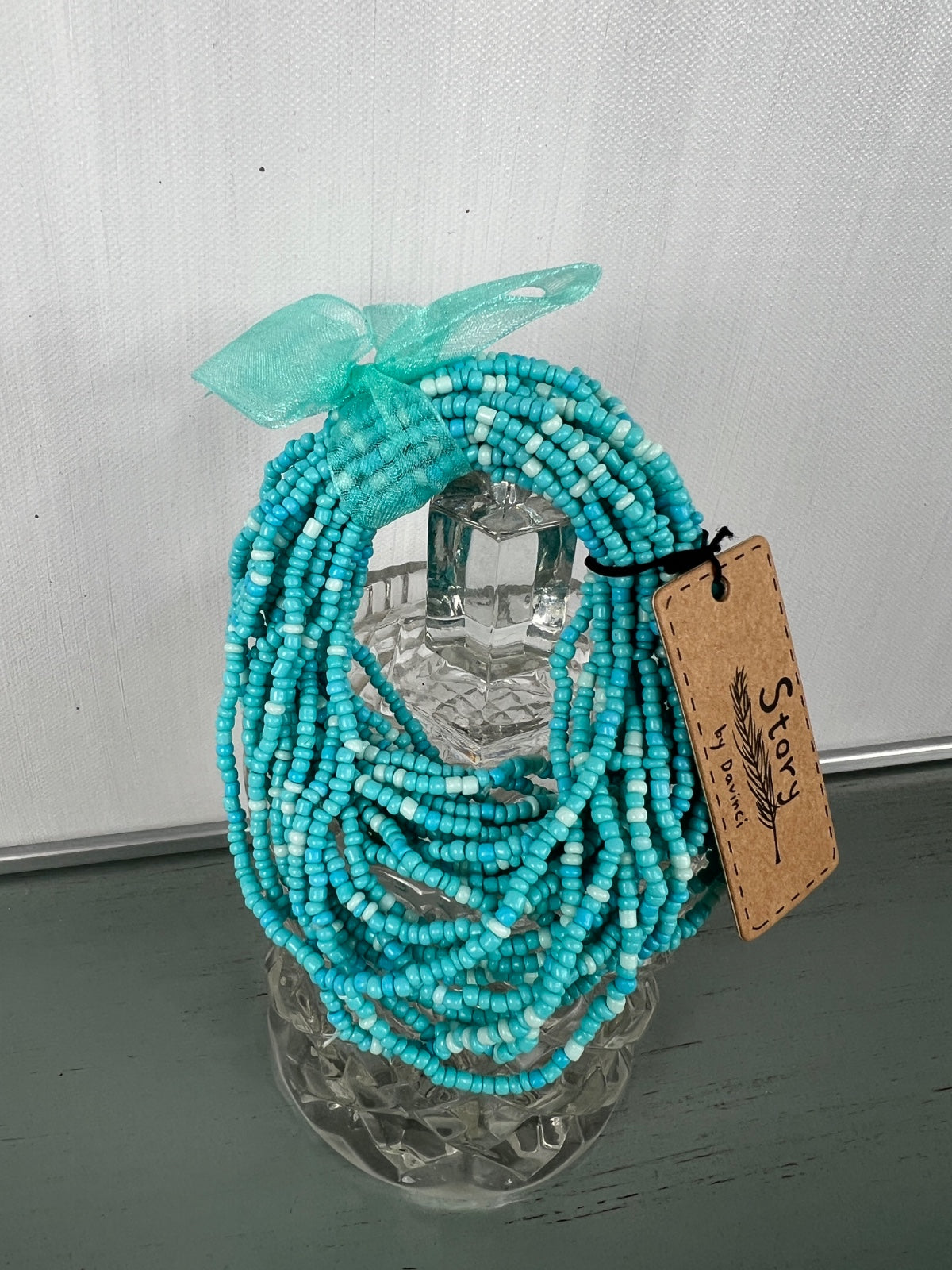 Turquoise beaded necklace with multiple strands of small seed beads arranged in a draped style, topped with a matching turquoise ribbon bow, displayed on a clear acrylic stand with a kraft paper price tag.