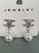 Pair of silver-tone drop earrings featuring ornate CC-shaped studs embellished with crystals, each suspending a white pearl. The earrings are displayed on a white jewelry card.
