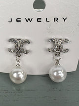 Pair of silver-tone drop earrings featuring ornate CC-shaped studs embellished with crystals, each suspending a white pearl. The earrings are displayed on a white jewelry card.