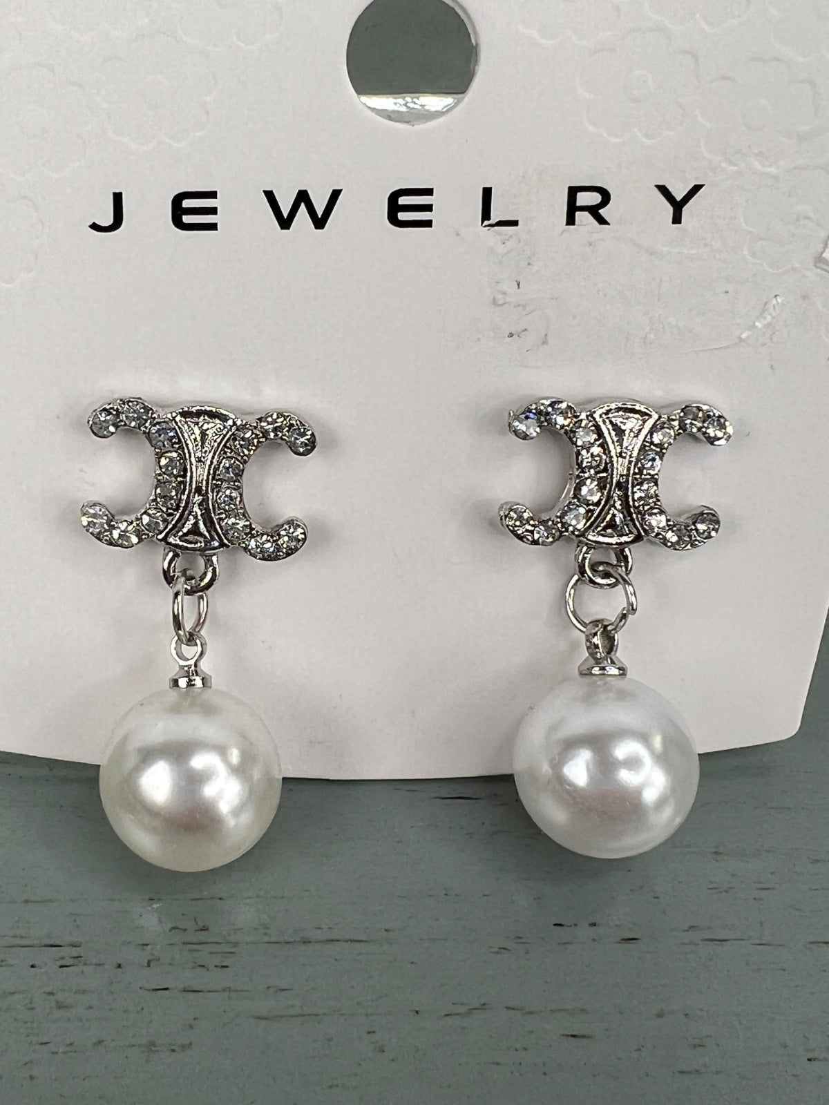 Pair of silver-tone drop earrings featuring ornate CC-shaped studs embellished with crystals, each suspending a white pearl. The earrings are displayed on a white jewelry card.