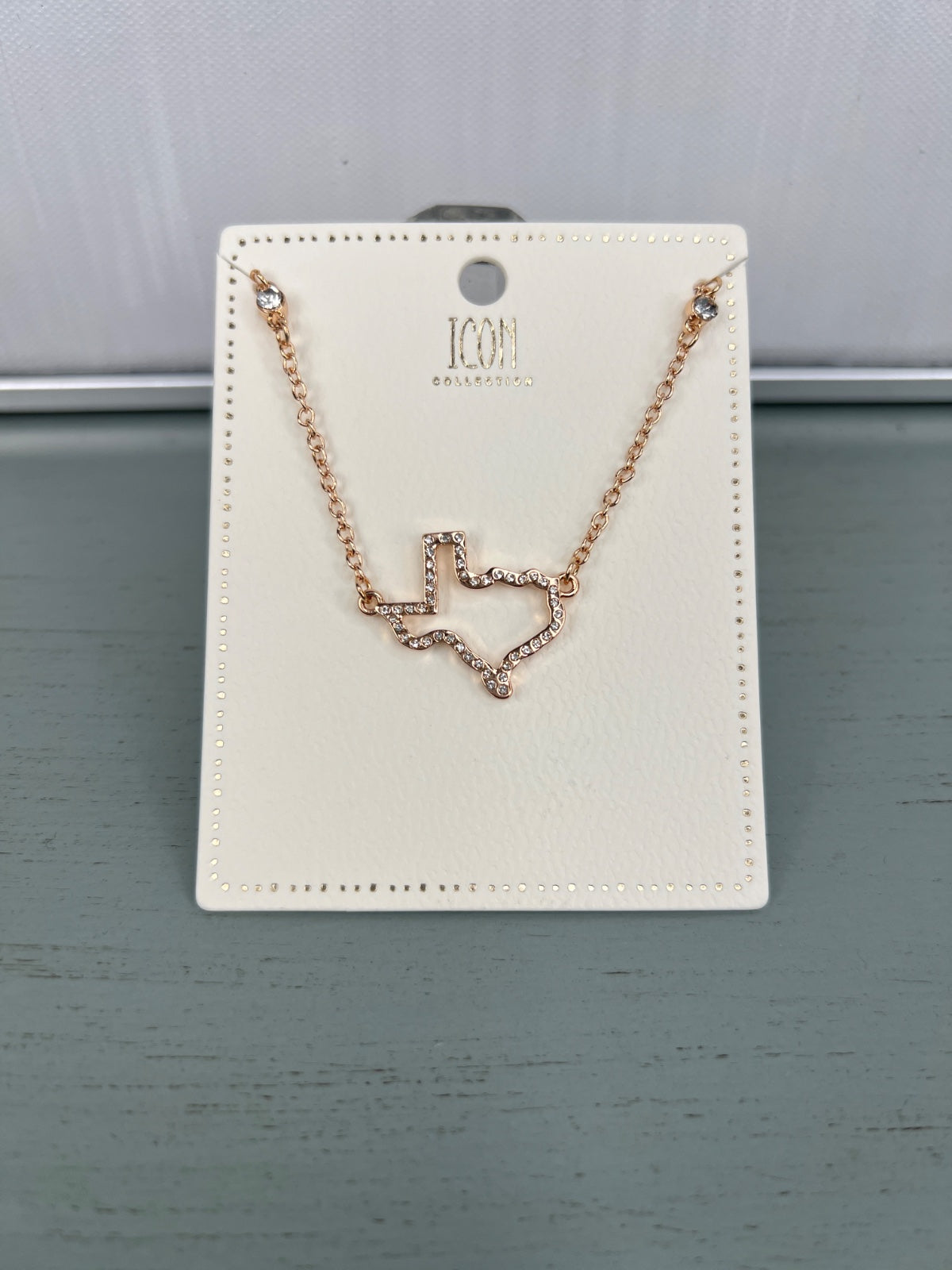 Rose gold-toned necklace with a delicate wavy heart pendant outlined in small crystals, displayed on a cream-colored rectangular jewelry card with decorative perforated border.
