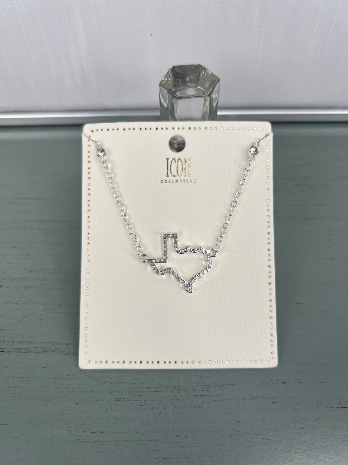 A silver-toned delicate chain necklace featuring a geometric V-shaped pendant with crystal embellishments, displayed on an ivory ICON branded jewelry card with decorative beading around the edges.