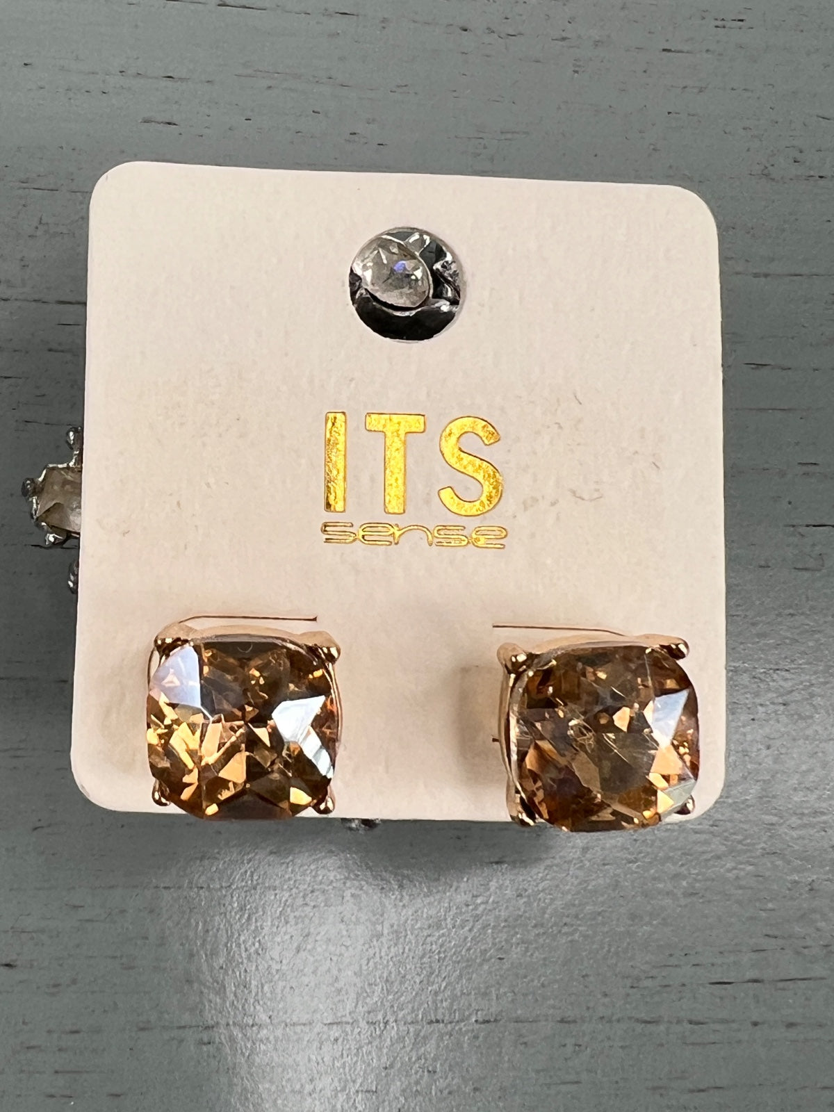 A pair of rose gold-toned stud earrings featuring large square-cut champagne or golden-brown crystals, displayed on a cream-colored ITS brand card with matching crystal accent studs shown above.