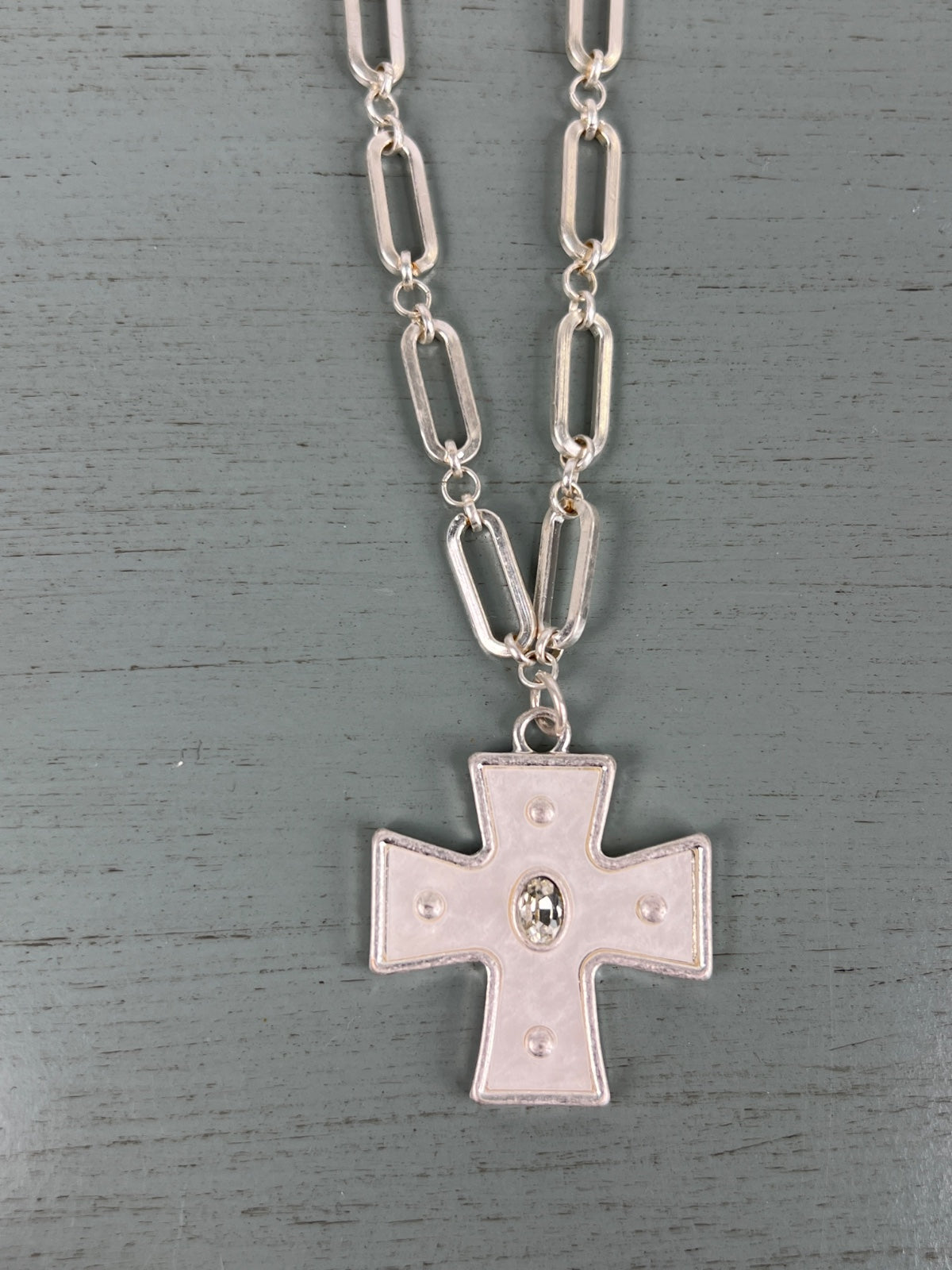 A silver-toned chain necklace featuring oval link segments, suspending a rectangular cross pendant with decorative dot details and a central oval medallion, displayed on a gray wooden background.
